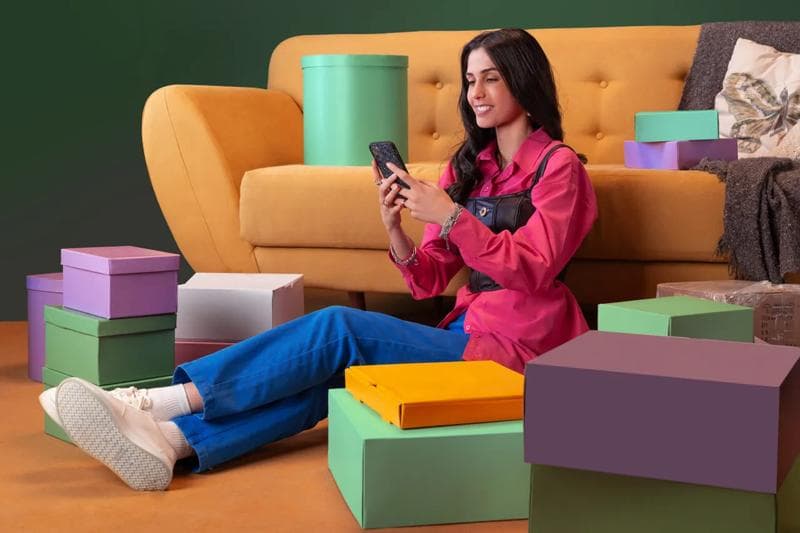 Woman with boxes - Woman sitting on the floor by a yellow sofa using a smartphone, surrounded by colorful Tabby delivery boxes.
