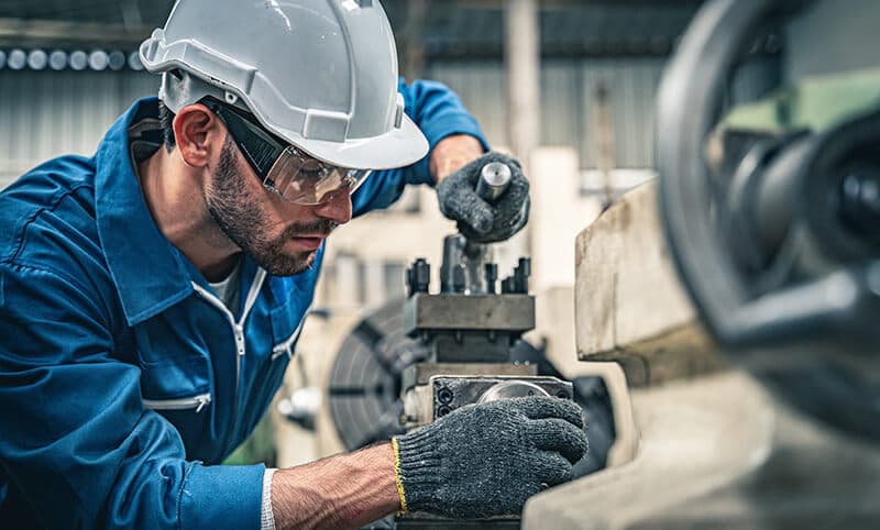Industrial worker wearing a white hard hat and safety glasses operating a precision metal lathe machine.