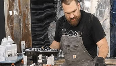 A man in grey overalls applying epoxy coating to a surface, with marbled stone-effect wall panels in the background.