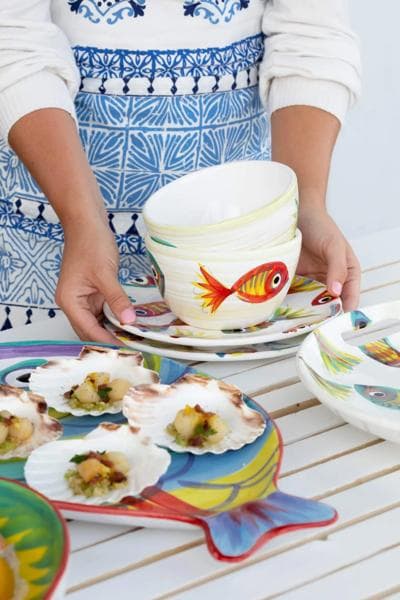 Hands holding a stack of Vietri fish-themed ceramic bowls and plates on a white outdoor table.