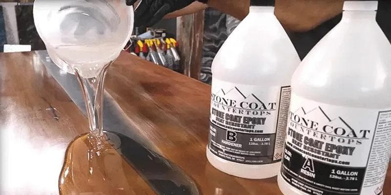 Stone Coat Countertops - Pouring clear Stone Coat Countertops epoxy resin onto a wood surface next to gallon bottles of resin and hardener.