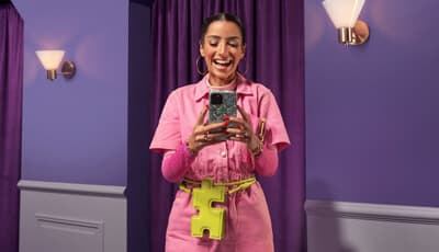 A smiling woman in a pink jumpsuit using a smartphone in a purple room with decorative wall sconces and a yellow puzzle-piece-shaped belt bag.