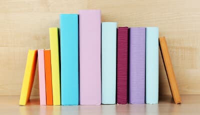 A row of books with colorful covers in shades of yellow, pink, orange, blue, and purple standing on a light wood shelf.