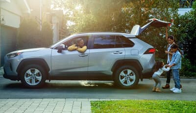 A family loading the trunk of a silver Toyota RAV4 SUV in a sunlit suburban driveway.