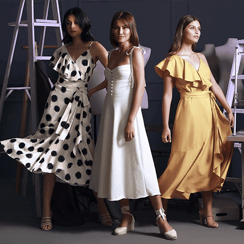 Three models wearing dresses in a sewing studio: one in a white and black polka dot wrap dress, one in a white midi dress, and one in a yellow ruffled dress.